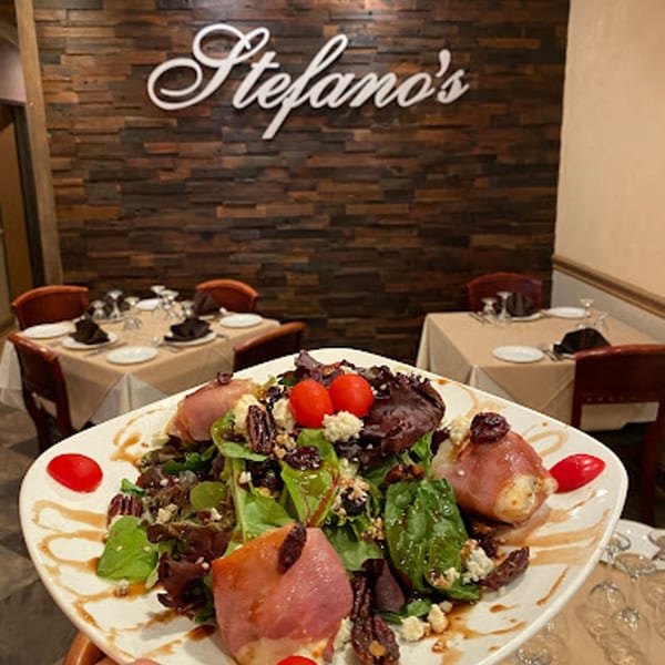 A fresh salad with prosciutto-wrapped cheese, greens, nuts, and tomatoes, with the Stefano's sign in the background.