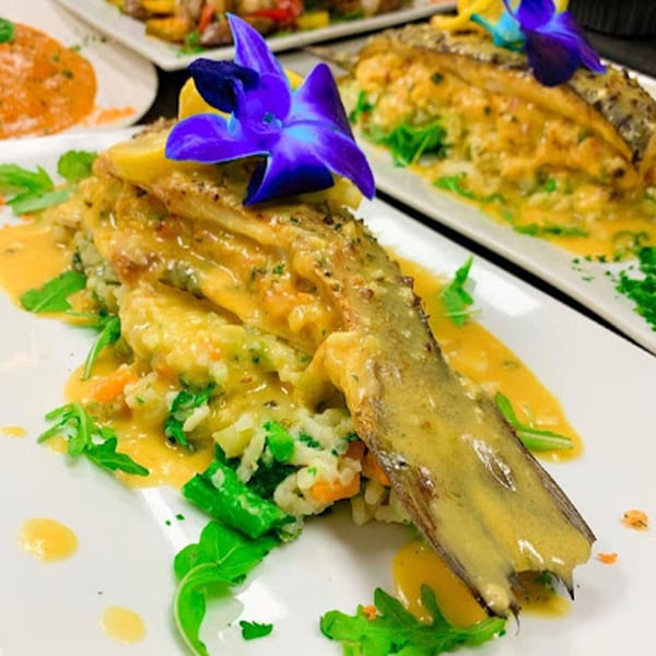 Plated fish entree with creamy sauce, risotto, and greens, garnished with a purple orchid.