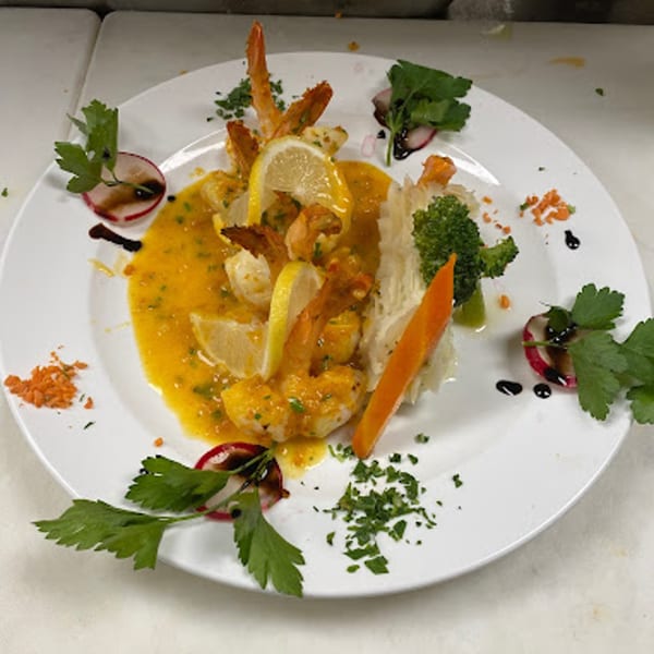 Shrimp scampi with lemon slices, broccoli, carrots, and radish garnishes on a white plate.